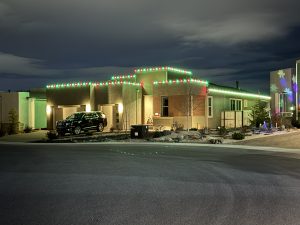 Night view of a modern house with green and red holiday lights outlining the roof, parked SUV in the driveway, cloudy sky above.